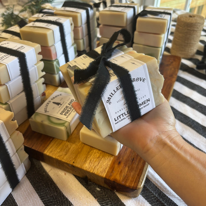 A hand holds a bar of handmade soap wrapped in a black ribbon and a white paper label
