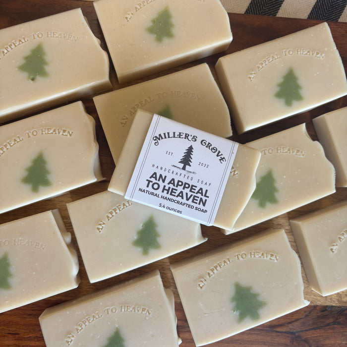 Multiple rectangular bars of handmade soap arranged on a wooden surface, with one bar centered and wrapped in a paper label that reads 'Smiler's Grove Handcrafted Soap An Appeal To Heaven.' Each soap bar is light cream with a small green tree motif and embossed text.