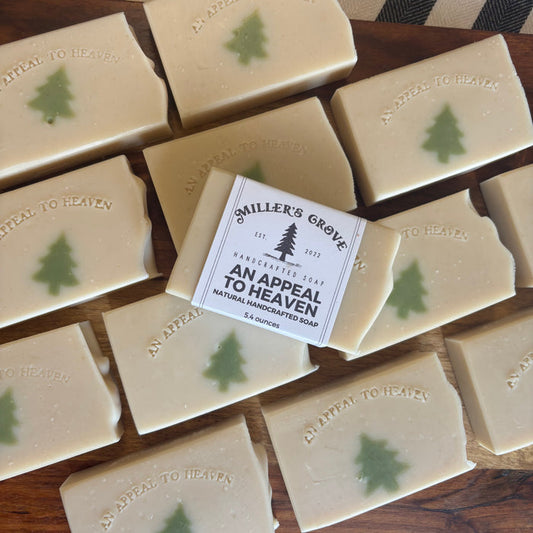 Multiple rectangular bars of handmade soap arranged on a wooden surface, with one bar centered and wrapped in a paper label that reads 'Smiler's Grove Handcrafted Soap An Appeal To Heaven.' Each soap bar is light cream with a small green tree motif and embossed text.