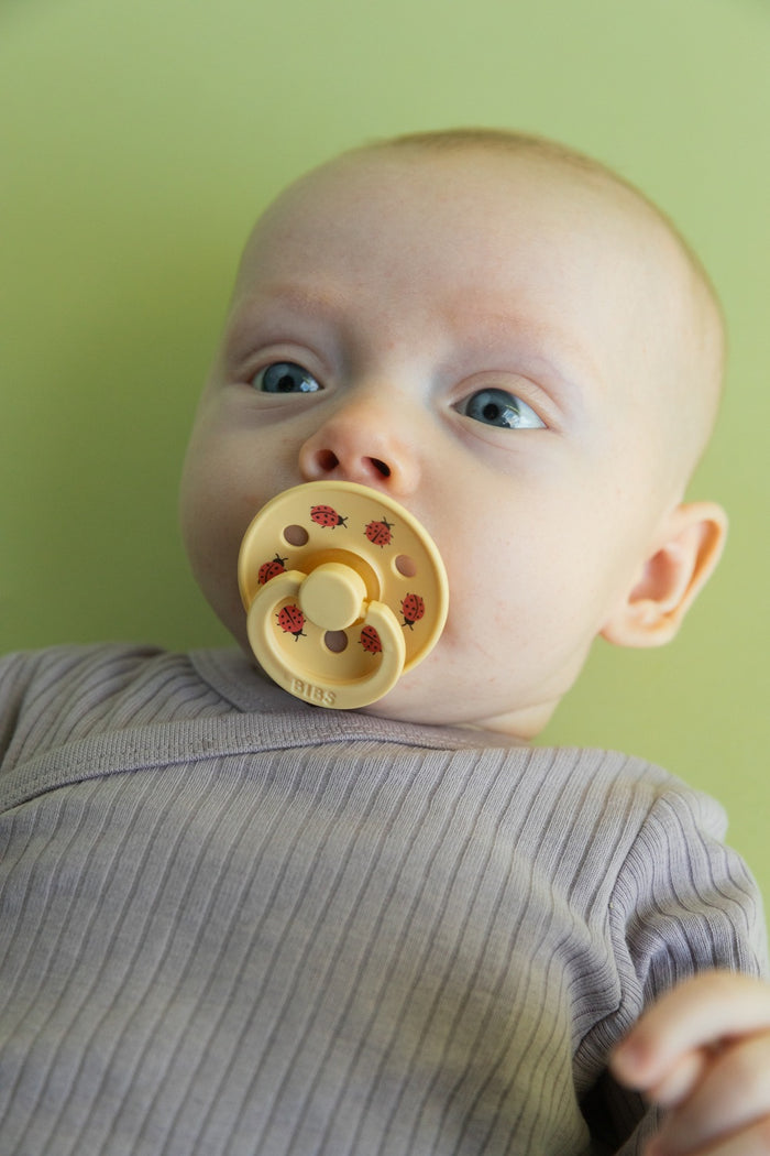Baby using BIBS Studio Colour Ladybug pacifier in pale yellow with red ladybug print, green background.