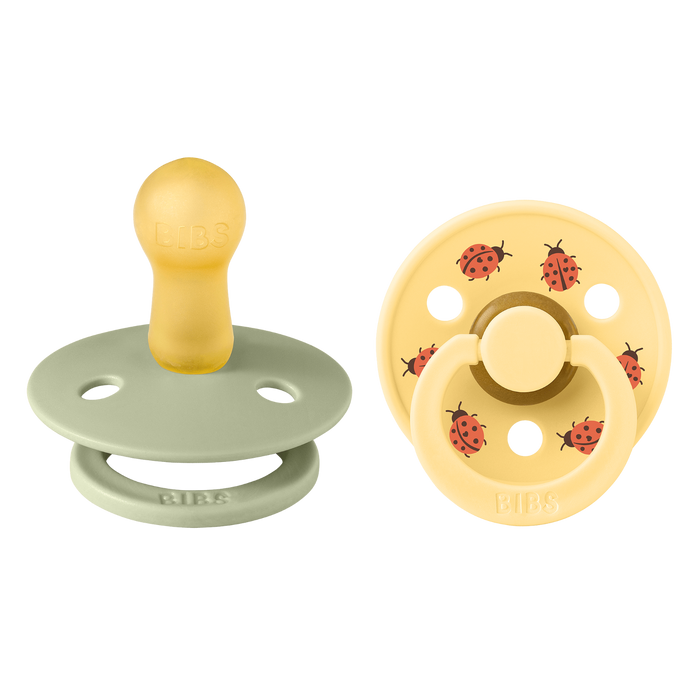 BIBS Studio Colour 2 Pack Ladybug pacifiers on white background: sage green and yellow with red ladybug prints, BIBS text on shields.