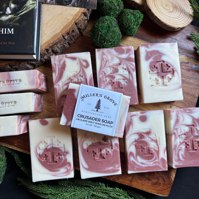 Assorted soap bars with a brand label on a wooden board, surrounded by greenery.