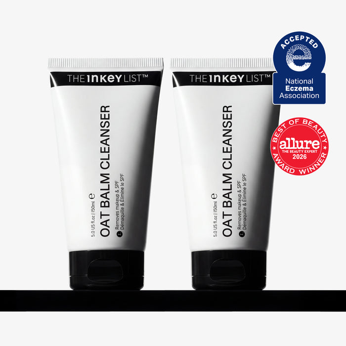 Two white squeeze tubes of THE INKEY LIST OAT Balm Cleanser with black caps, displayed side-by-side on a black surface against a white background, with two award badges on the right.